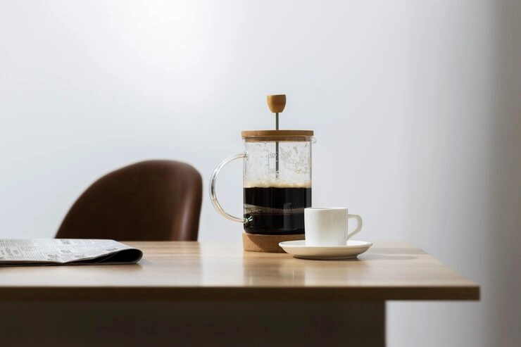 A French press with coffee, a white cup and saucer, and a newspaper on a wooden table