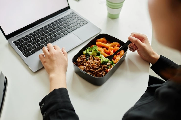 woman eating takeout meal with noodles shrimp and broccoli while working on laptop at desk